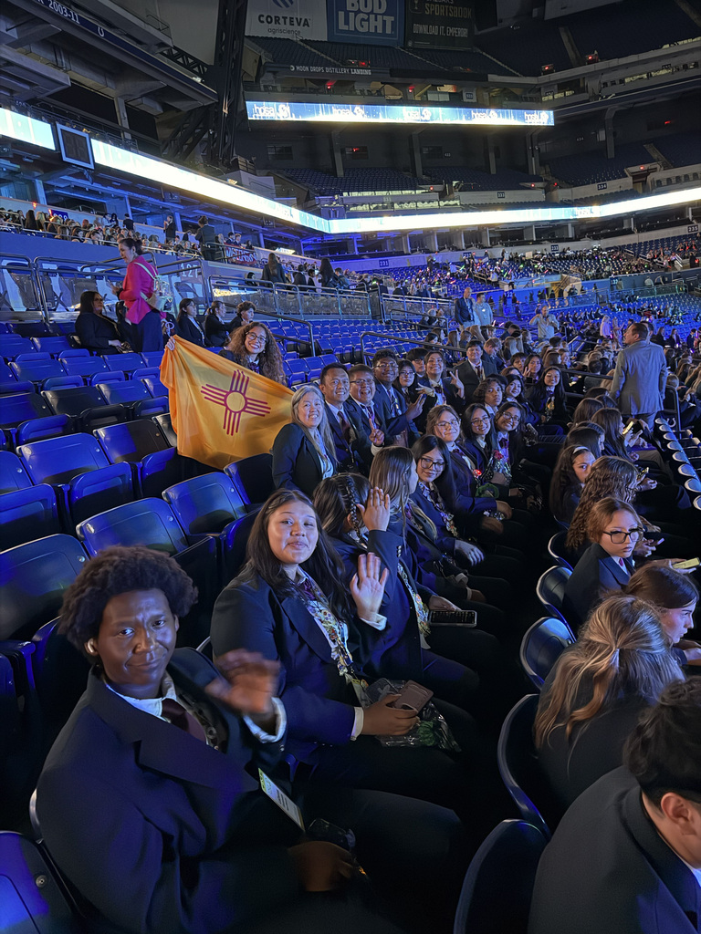 Large crowd of people in blue seats at an event. Yellow flag with design. Bright overhead lighting.