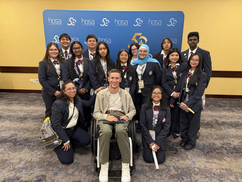 Group of students and a man in a wheelchair pose for a photo in front of a blue banner.