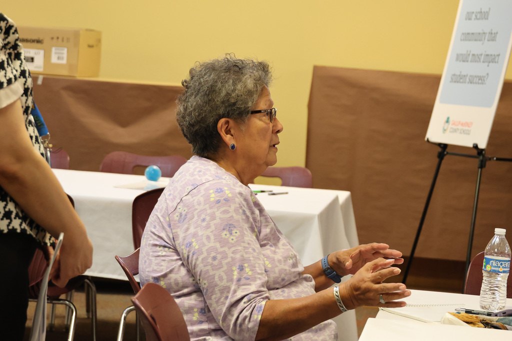 An older woman in a floral shirt sits at a table in a meeting, gesturing with hands.