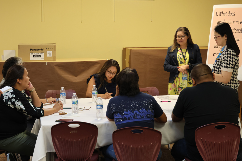 Group of people seated around a table in a room with yellow walls. One person stands at the head of the table, addressing others.