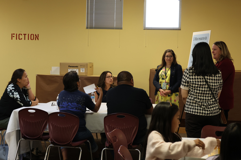 Group of adults seated around a table with papers, one woman presenting at a board.