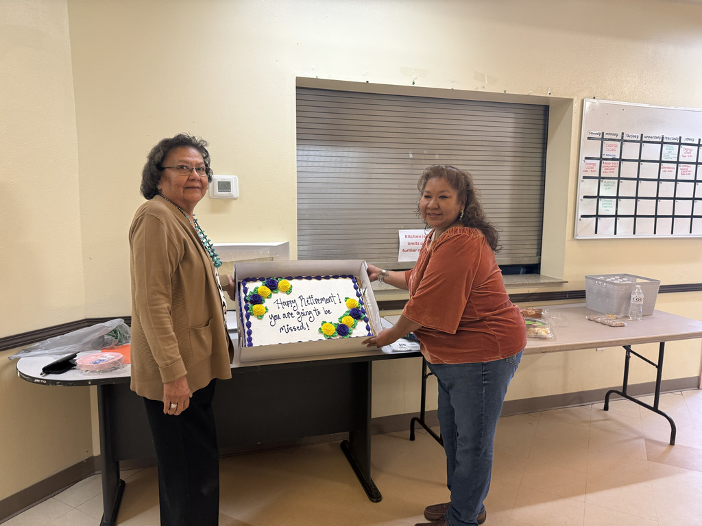 Two women stand beside a table, holding a cake box with a colorful design. Behind them is a window with blinds.