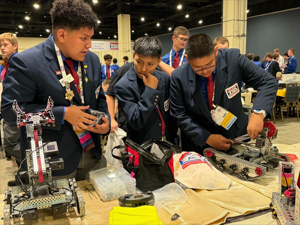 Three students in suits and ties work on a table with robotic equipment and parts.