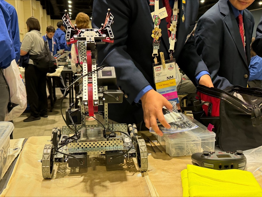 People in suits examine a robot on a table, with more people and tables in the background.