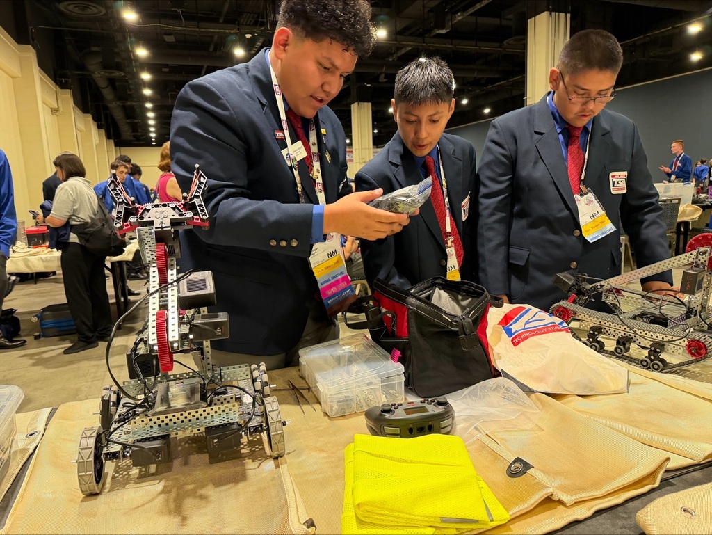 Three students in suits examine a mechanical robot on a table in a large hall.