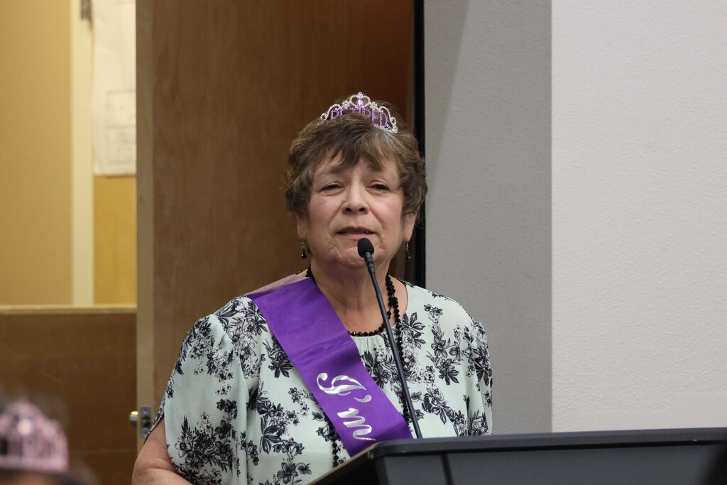 A woman in a floral shirt and purple sash stands at a podium, wearing a tiara. She is speaking into a microphone.