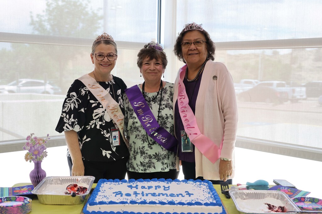 Three women wearing sashes stand by a cake. A table displays the cake and various containers.