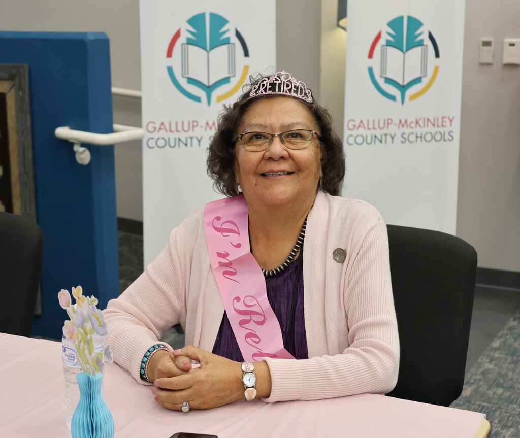Woman wearing a tiara and sash with "Miss Kei" written on it sits at a table.