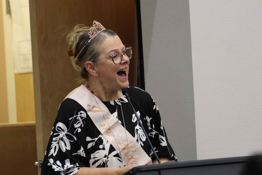Woman speaking into a microphone wearing glasses and a sash that reads "Sr. Nurse".