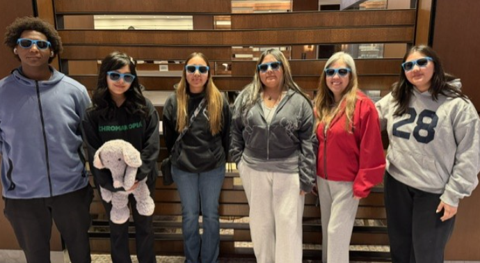 A group of people wearing sunglasses stand together in front of a wooden wall.