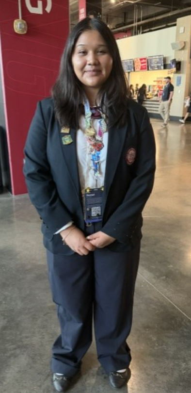 A woman in a navy suit with a white shirt and colorful lanyard stands in an indoor hall.