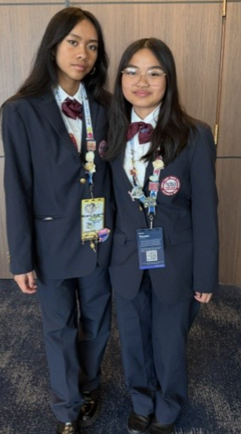 Two individuals in navy blue suits with bow ties and lanyards stand in a corridor.