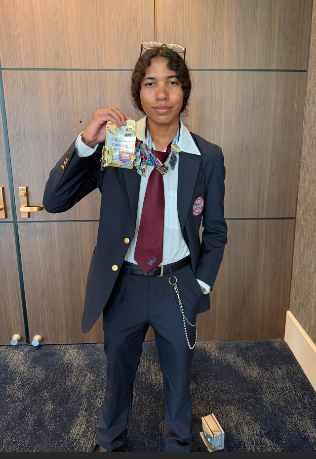 Person in a suit holding a medal and standing in front of a wood-paneled wall.