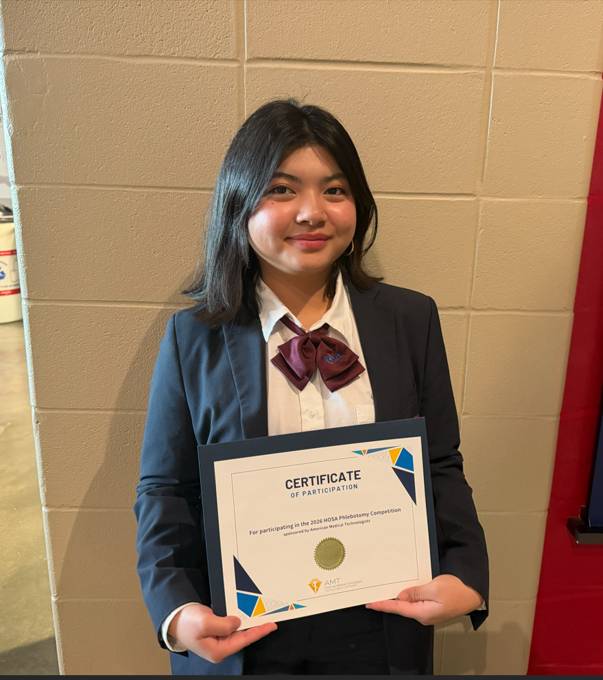 A young person holds a certificate with a smiling face. They are wearing a black suit with a bow tie.