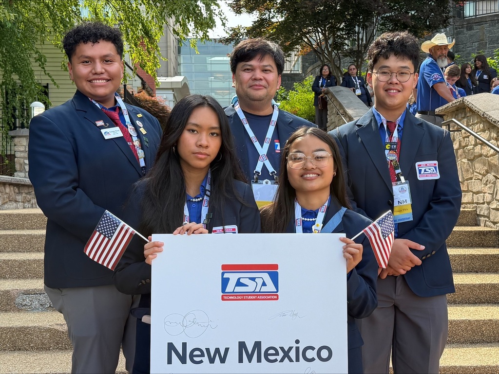 Five people in suits stand on stairs, holding a sign that says "New Mexico" and flags.