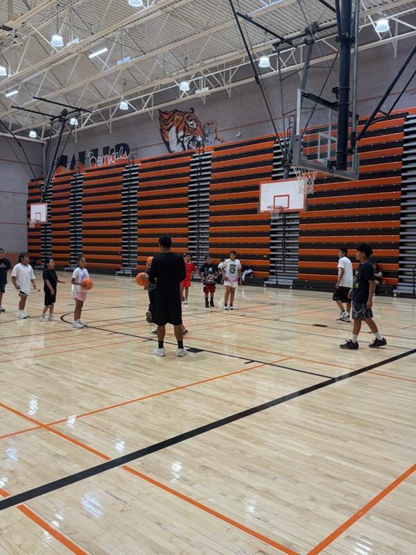 An indoor basketball court with several people standing. Players hold basketballs and stand in groups. The court has wooden floors and orange markings.