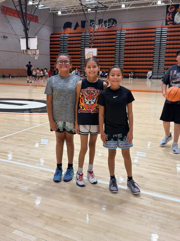 Three young girls pose for a photo on a basketball court. Two wear sneakers, one with glasses. A basketball hoop and bleachers are in the background.