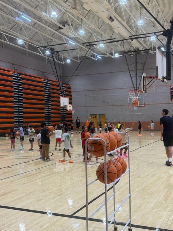 A basketball court with people, some holding basketballs, and a rack with several balls.