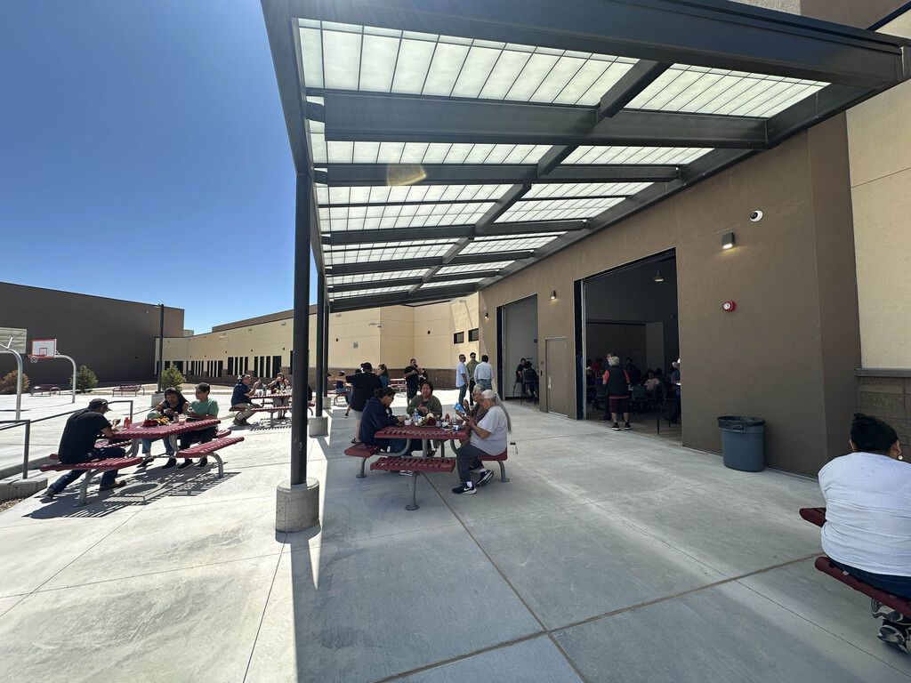 People sit at tables under a metal canopy on a sunny day. The area is paved, with a building in the background.