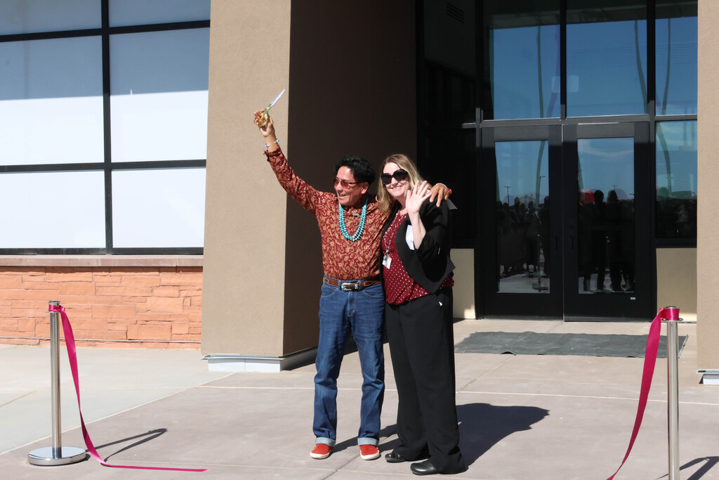Two people stand next to each other, with one raising a trophy, outside a building with glass windows.