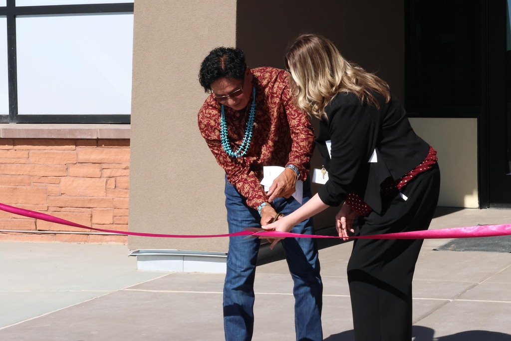 Two people in a building entranceway cut a pink ribbon as part of a ceremony.