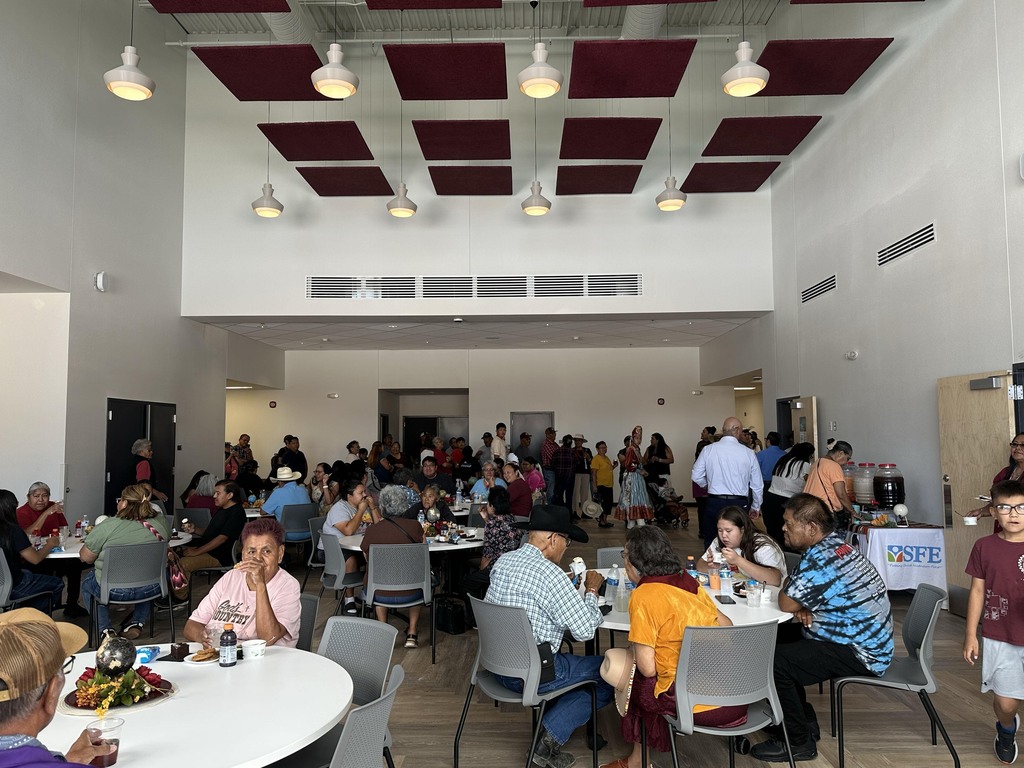 A dining area with tables and chairs, a group of people sitting and standing, a white ceiling with lights, and a sign on a table.