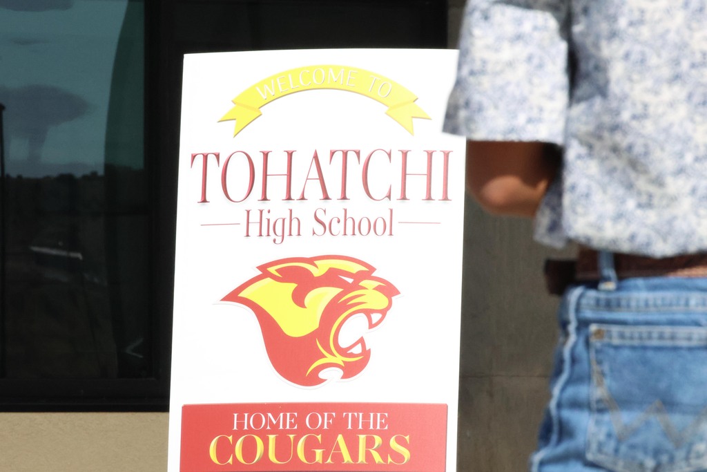 A sign for Tohatchi High School shows a yellow and red cougar, with a person in jeans standing nearby.