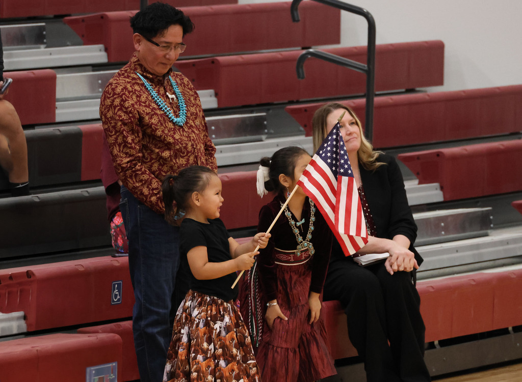 A man and two children, one holding an American flag, stand on a bleacher. A woman in black sits on the bench behind them.