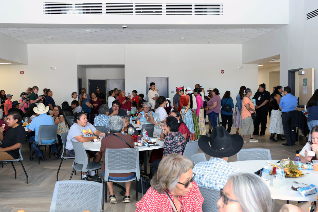 A large group of people gathered in a room with tables and chairs. Some wear hats and glasses.