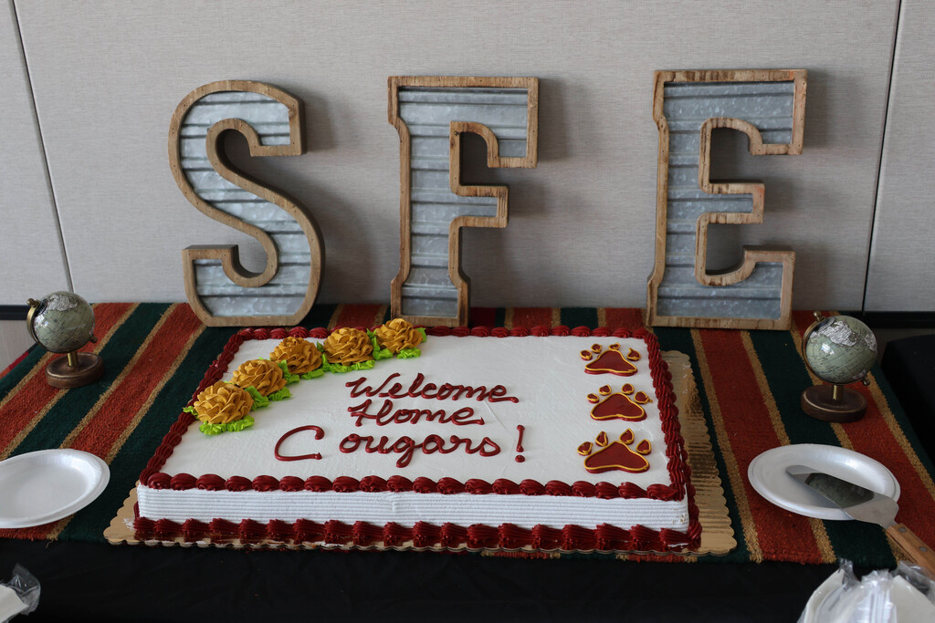 A large cake with red and white icing displays "Welcome Home, Cougars!" in red letters. Behind it, large letters spell "SFE."