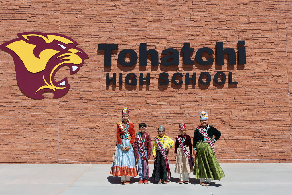 Five people in traditional attire stand before a brick wall. A logo is above them. Text reads "Tohatchi High School".