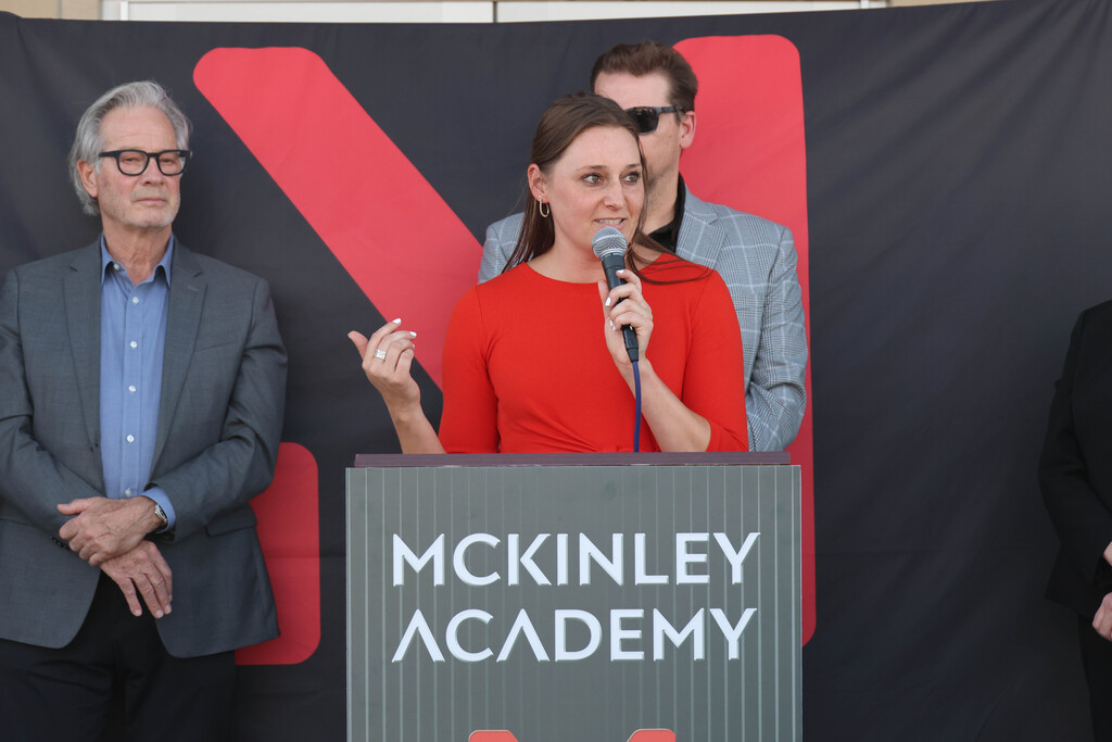 A woman speaks at a podium with "McKINLEY ACADEMY" displayed. Two men in suits stand behind her.