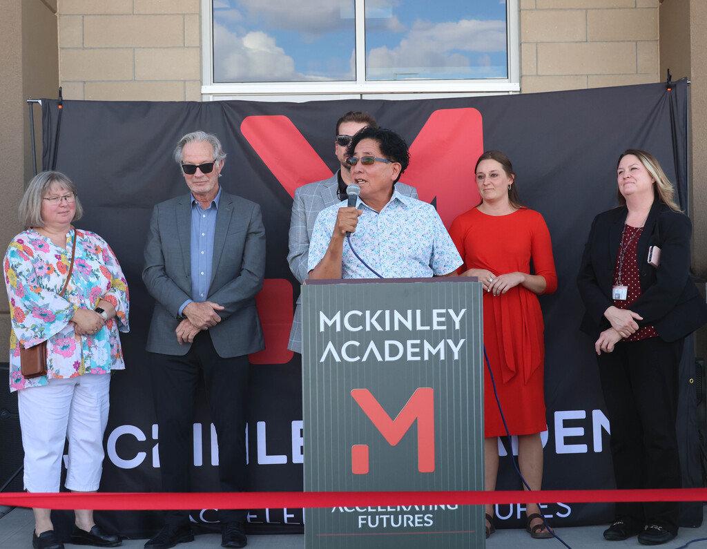 A group of adults stand behind a podium with a McKinley Academy banner. A man speaks at the podium.