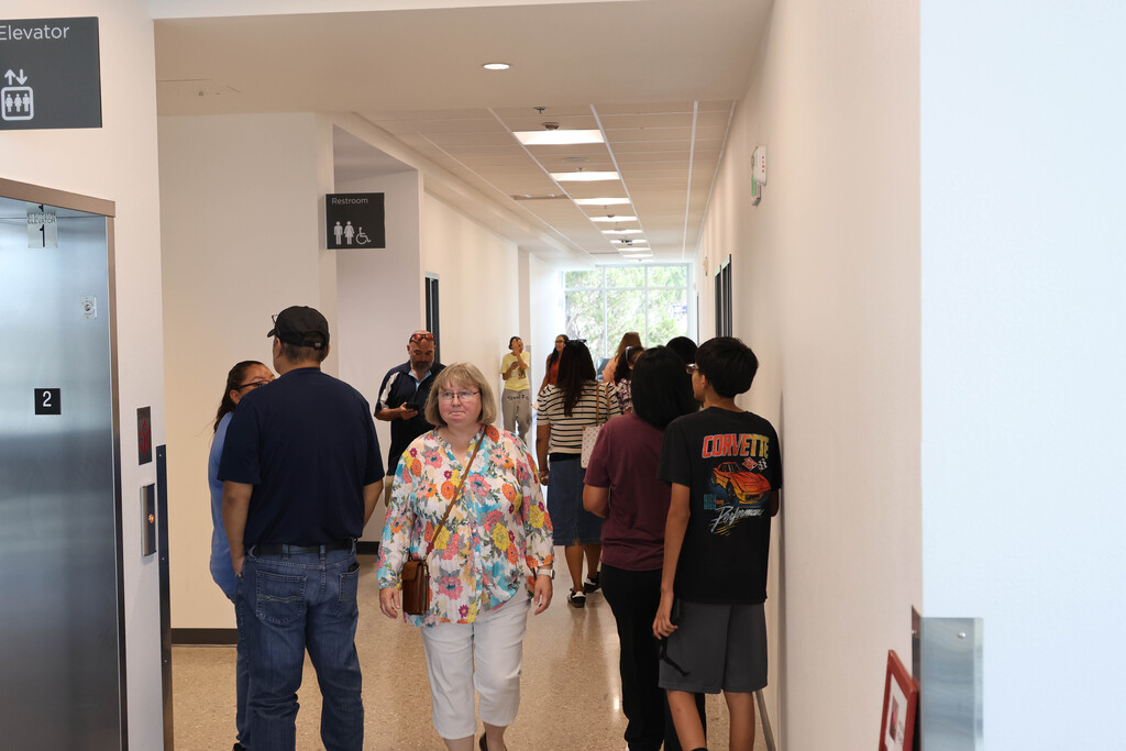 A group of people in a hallway with white walls, ceiling lights, and an elevator on the left.