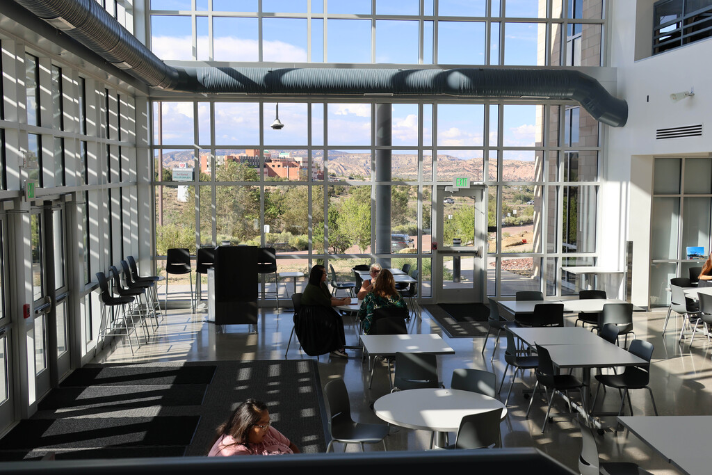 A modern interior with glass walls and tables. Several people sit at tables. Sunlight shines through.