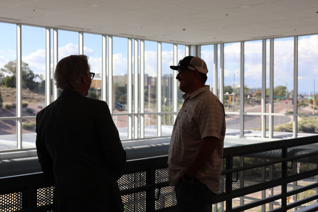 Two men in hats and glasses stand on a balcony, facing each other. Large windows show a view of a road.