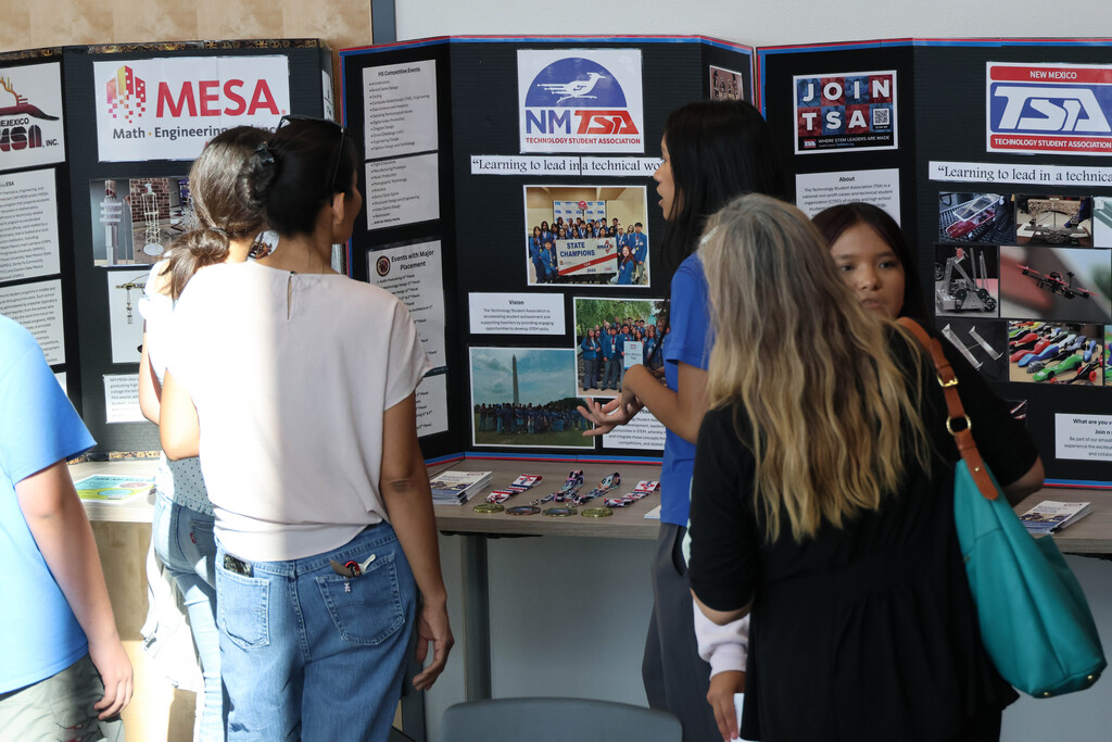 People gather around a display board with logos and images, such as NM TSA, with several individuals examining the contents.
