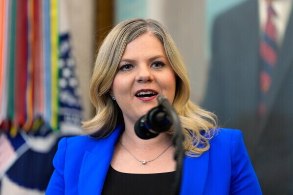 A woman with blonde hair speaks into a microphone. She wears a blue blazer and black top. Behind her, flags and a man's portrait are visible.