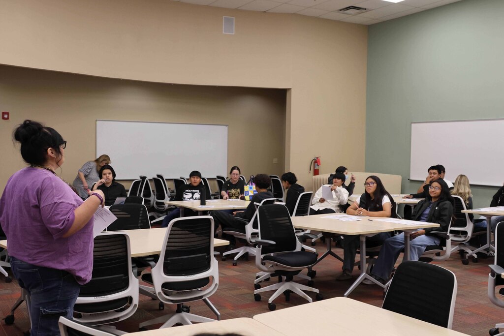 A woman stands near a whiteboard, addressing a group seated at tables in a classroom.