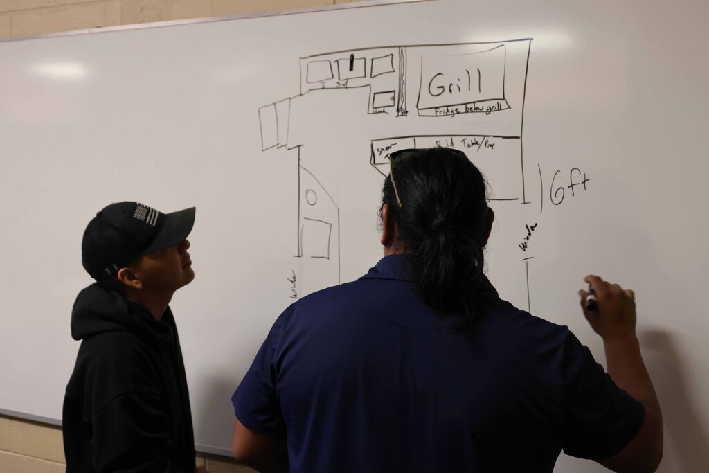 Two individuals look at a whiteboard with a building layout, one pointing with a marker.