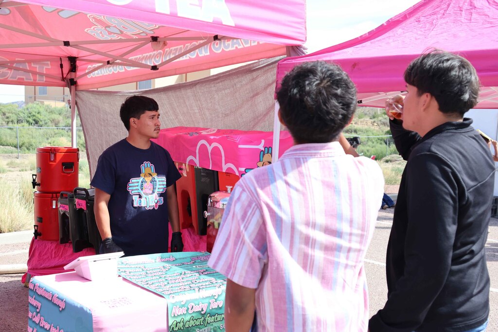 Three people gather under a pink canopy. Two speak while the third holds a drink. A table holds a colorful design.