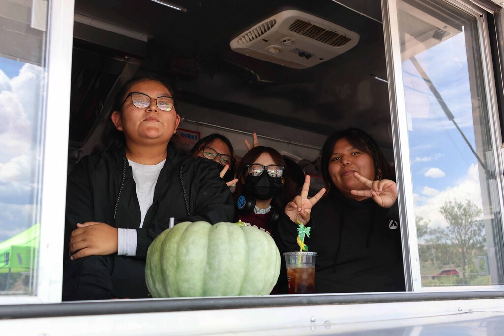 Four individuals inside a food truck, two making peace signs. A large green gourd is between them.