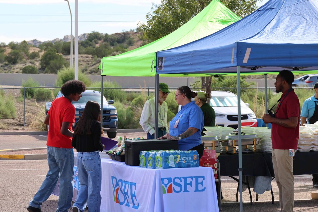 People gather under tents with tables, one blue, one green, and another white. They stand and walk in a parking area.