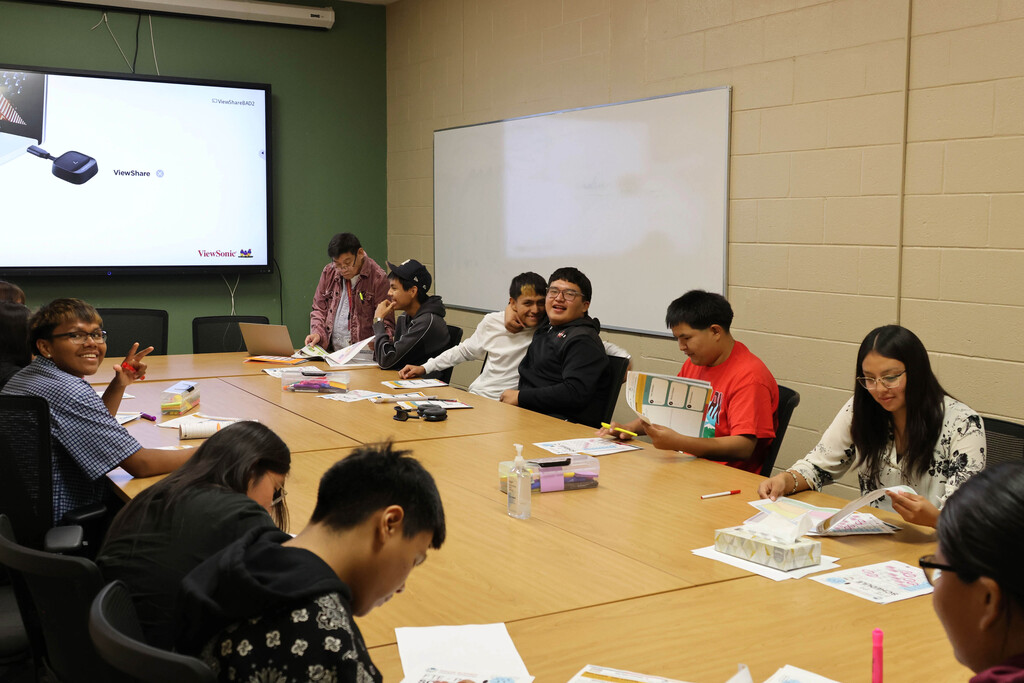 A classroom with participants seated around a table. A projector screen displays content. Whiteboard in background.
