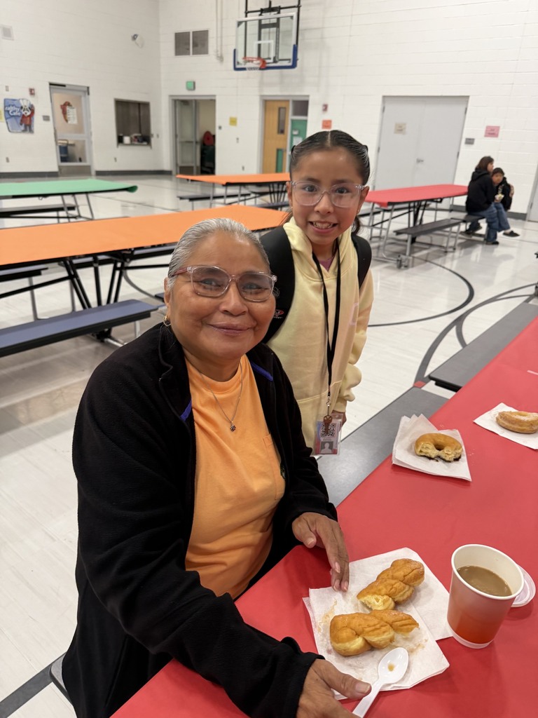 Two people sit at a table in a cafeteria, smiling. A woman in glasses wears a black jacket.