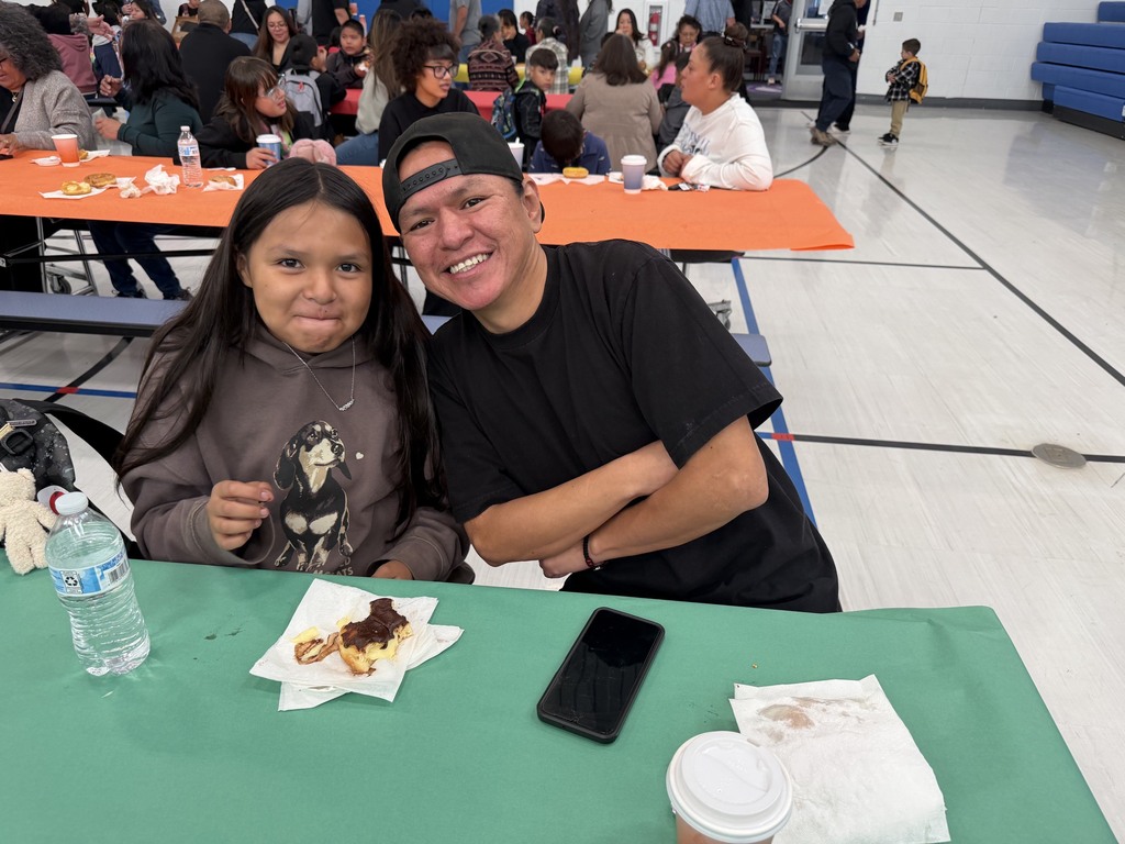 A smiling adult and a child sit at a table in a gym. They have food and a phone.