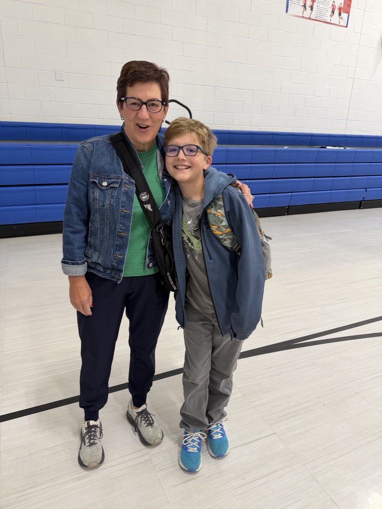 An adult and a child stand together in a gym. The adult wears glasses, a jacket, and sneakers. The child wears glasses and a jacket.