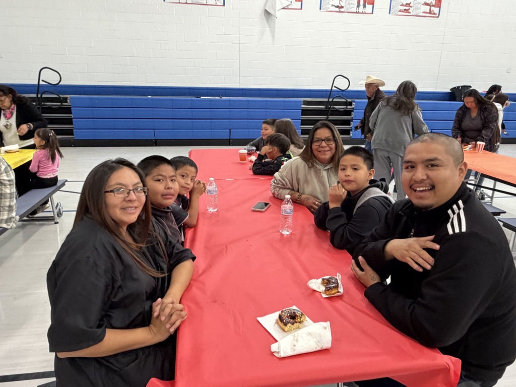 A family sits at a long red table in a gym with blue bleachers, smiling for a picture.