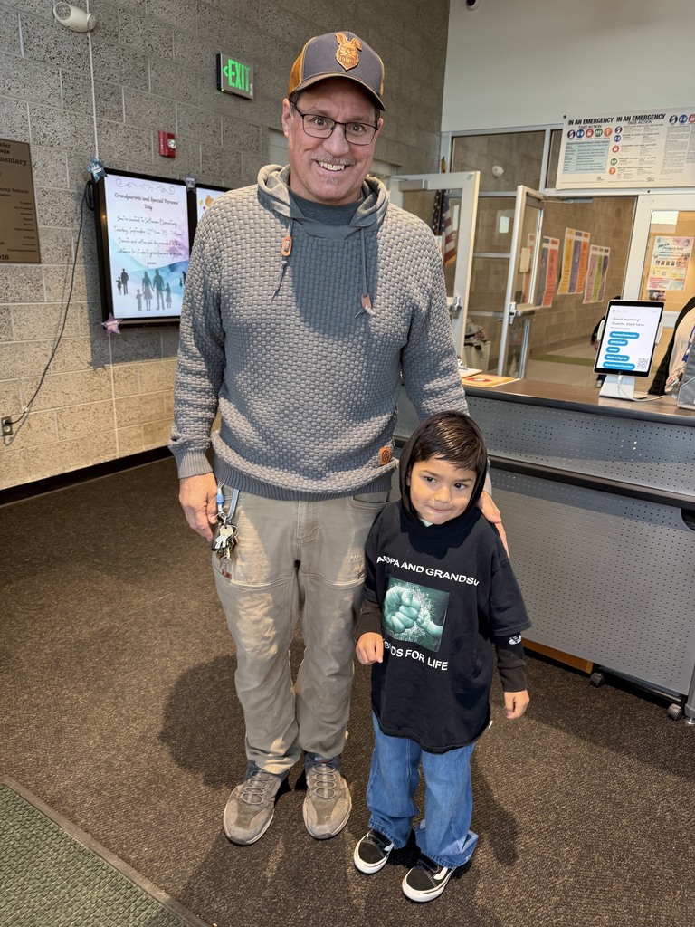 A man and a child stand in a hallway. The man wears a hat, glasses, and a gray hoodie. The child wears a black sweatshirt.