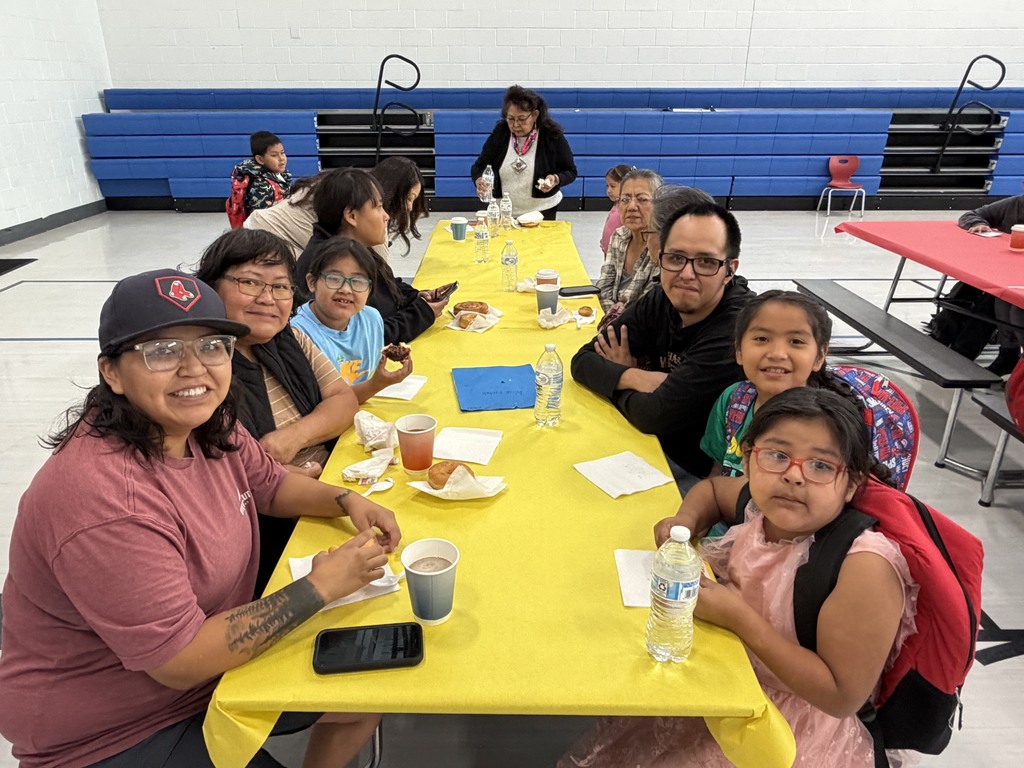 Group of people seated at a table, smiling, with plates, cups, and water bottles. Tables have yellow cloths. Background features blue walls and bleachers.